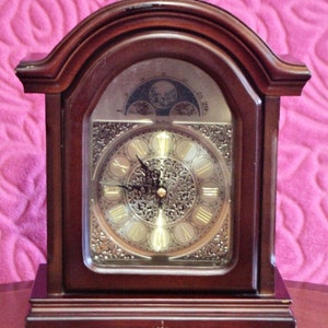 Puede incluir: Un reloj de chimenea de madera oscura con una esfera dorada y marcadores de hora con números romanos. El reloj tiene un diseño decorativo alrededor de la esfera y una pantalla de fase lunar en la parte superior.