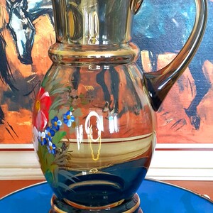 May include: A dark blue glass jug with a gold rim and painted floral design. The jug has a wide mouth and a curved handle.