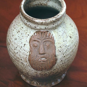 Può includere: Un vaso in ceramica macchiata con corpo rotondo e piccolo manico. Il vaso presenta un volto scolpito con la barba. I colori sono bianco sporco, marrone e marrone scuro. Il vaso è un oggetto decorativo.