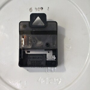 May include: Close-up of the back of a clock mechanism with a black plastic casing. The mechanism is labeled "+1ECL LR 1A" and "MADE IN GERMANY".