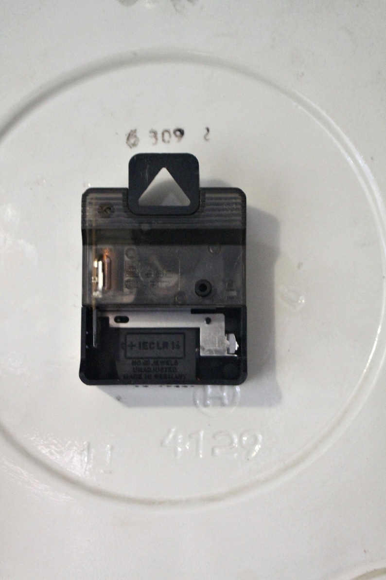 May include: Close-up of the back of a clock mechanism with a black plastic casing. The mechanism is labeled "+1ECL LR 1A" and "MADE IN GERMANY".