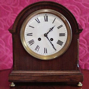 Puede incluir: Un reloj de chimenea de madera oscura con una esfera blanca y marcadores de hora romanos negros. El reloj tiene un borde de latón alrededor de la esfera y un péndulo de color dorado.