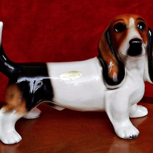 Könnte beinhalten: Eine Keramikfigur eines braunen, schwarzen und weißen Basset-Hundes. Die Figur steht auf einer Holzoberfläche. Der Hund hat eine weiße Brust und einen schwarzen und braunen Rücken. Die Figur ist auf der Unterseite mit "Made in England" gekennzeichnet.