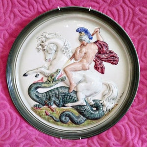 Puede incluir: Un plato de cerámica blanco con un diseño en relieve de un guerrero a caballo matando a un dragón. El guerrero lleva un casco y una capa roja. El caballo es blanco y el dragón es verde.