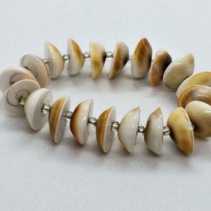 Pode incluir: Um bracelete feito de pequenas conchas brancas e marrons enfiadas em contas prateadas.