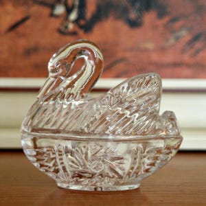 May include: A clear glass swan-shaped trinket box with a detailed, cut-glass design. The box is closed and has a decorative, floral pattern on the base.