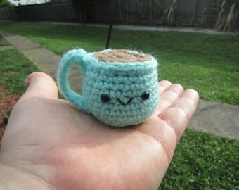 May include: A small crocheted mug with a brown crocheted coffee or cocoa inside. The mug has a light blue color and a happy face with black eyes.