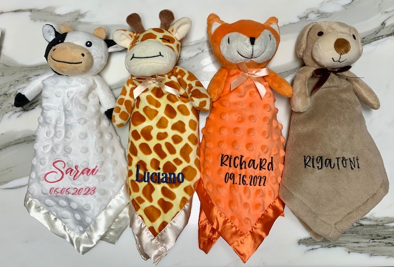 Peut inclure: Quatre couvertures pour b&eacute;b&eacute; en peluche avec des animaux et des noms brod&eacute;s ainsi que des dates. Les couvertures sont blanches, brunes, orange et beige. Les animaux sont une vache, une girafe, un renard et un chien.