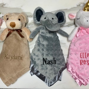 Peut inclure: Trois couvertures pour b&eacute;b&eacute; personnalis&eacute;es avec des t&ecirc;tes d'animaux en peluche. Les couvertures sont beige, grise et rose avec des noms brod&eacute;s "Kaylani", "Nash" et "Ella Rose".