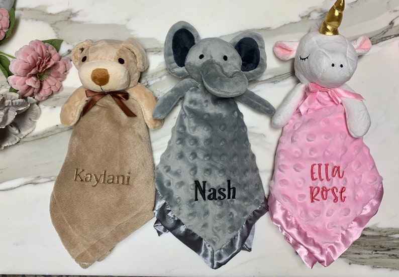 Peut inclure: Trois couvertures pour b&eacute;b&eacute; personnalis&eacute;es avec des t&ecirc;tes d'animaux en peluche. Les couvertures sont beige, grise et rose avec des noms brod&eacute;s "Kaylani", "Nash" et "Ella Rose".