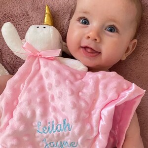 Peut inclure: Une couverture pour b&eacute;b&eacute; rose et blanche avec un jouet en peluche en forme de licorne blanche. La couverture a une bordure en satin et le nom "Leilah Jayne" brod&eacute; en fil bleu.