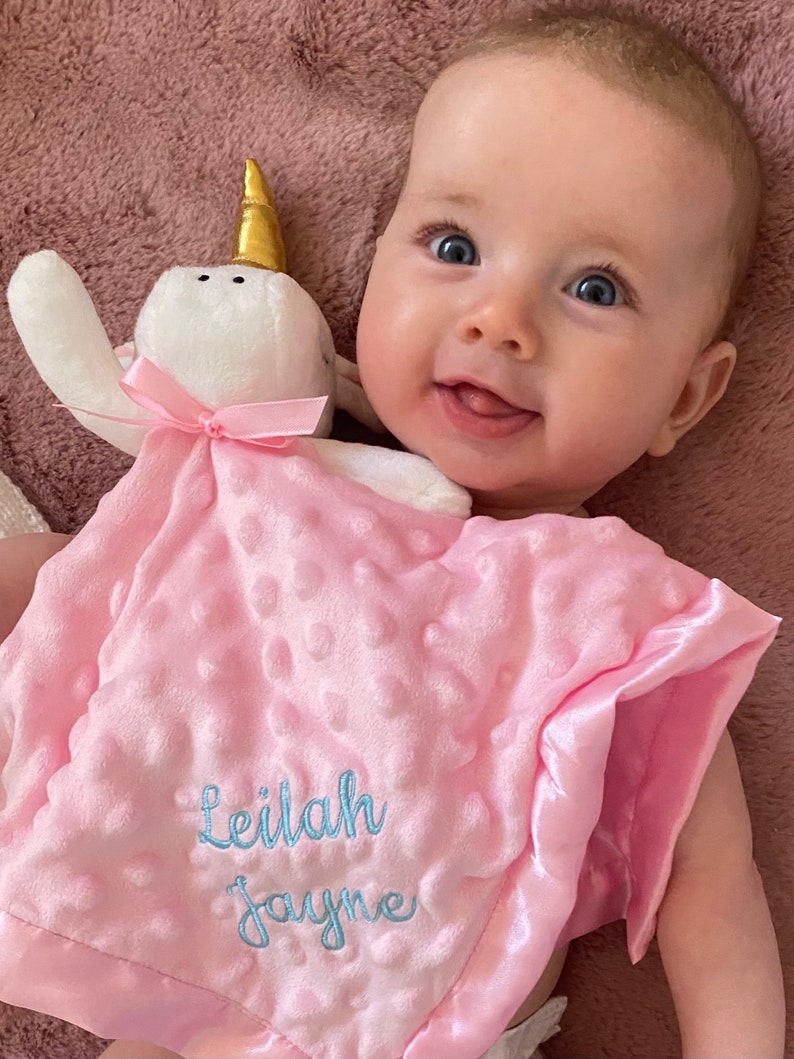 Peut inclure: Une couverture pour b&eacute;b&eacute; rose et blanche avec un jouet en peluche en forme de licorne blanche. La couverture a une bordure en satin et le nom "Leilah Jayne" brod&eacute; en fil bleu.