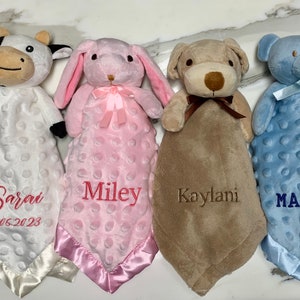 Peut inclure: Quatre couvertures pour b&eacute;b&eacute; personnalis&eacute;es avec des jouets en peluche attach&eacute;s. Les couvertures sont blanches, roses, brunes et bleues avec des noms brod&eacute;s "Sarai 06.05.2023", "Miley", "Kaylani" et "MATHEW".