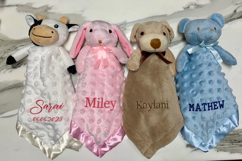Peut inclure: Quatre couvertures pour b&eacute;b&eacute; personnalis&eacute;es avec des jouets en peluche attach&eacute;s. Les couvertures sont blanches, roses, brunes et bleues avec des noms brod&eacute;s "Sarai 06.05.2023", "Miley", "Kaylani" et "MATHEW".