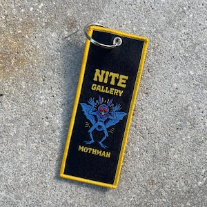 May include: Black key chain with yellow stitching featuring a blue mothman illustration and the text "NITE GALLERY" and "MOTHMAN".