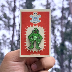 May include: A green enamel pin featuring a creature from the black lagoon, with a red background and the text "ENAMEL PIN". The pin is on a card with the text "NITE GALLERY" and a skull and crossbones design.