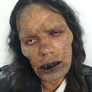 Peut inclure: Gros plan d'un visage de zombie avec une peau pâle, des cheveux foncés et des yeux jaunes. Le visage a une apparence de décomposition avec des dents exposées et des taches de sang. La personne porte une veste noire et une chemise blanche.