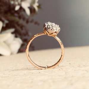 Könnte beinhalten: Ein goldener Ring mit einem Cluster aus kleinen weißen Steinen, die in der Mitte eingesetzt sind. Der Ring liegt auf einer hellbraunen Oberfläche.