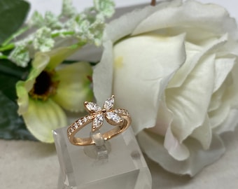 May include: A gold ring with a flower-shaped design, set with marquise-cut gemstones at the centre and smaller stones on the band. The ring is displayed on a clear stand, with white flowers in the background.