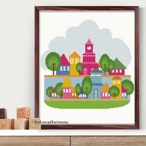 May include: A framed cross-stitch art piece depicting a colorful town scene. The artwork features houses in various colors, including pink, yellow, and blue, with green trees and a cloudy sky. The frame is a dark wood. The text "DeLoraiPatterns" is visible.