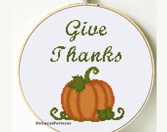 May include: A cross-stitch embroidery hoop featuring the words "Give Thanks" in green above an orange pumpkin with green leaves. The design is set against a white background, perfect for fall decor.