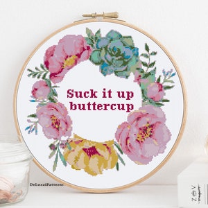 May include: A white embroidery hoop with a floral wreath design and the text "Suck it up buttercup". The wreath is made of pink and yellow flowers and green leaves.