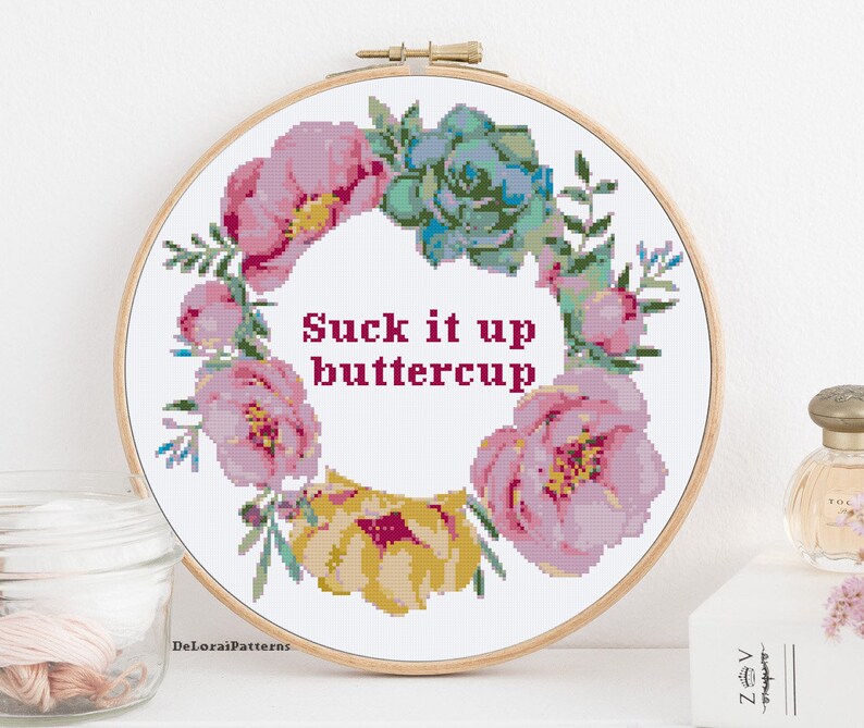 May include: A white embroidery hoop with a floral wreath design and the text "Suck it up buttercup". The wreath is made of pink and yellow flowers and green leaves.