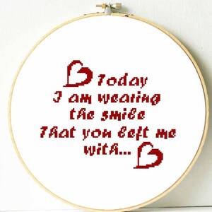 May include: A cross-stitch embroidery hoop with the text "Today I am wearing the smile that you left me with..." in red. The design includes two red pixelated hearts. The hoop is made of light-colored wood.