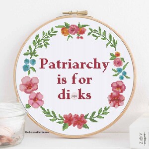 May include: A cross-stitch embroidery hoop with the text "Patriarchy is for dicks" in red. The text is surrounded by a floral wreath of pink, orange, and blue flowers and green leaves. The embroidery is on a white background.
