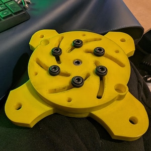 Puede incluir: Un componente mecánico amarillo brillante de varias capas con seis tornillos negros. El componente tiene un centro circular con ranuras curvas y cuatro extensiones redondeadas con agujeros. Se encuentra sobre una superficie oscura.