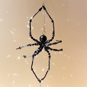 Könnte beinhalten: Ein schwarzes, perlenbesetztes Spinnenornament, das an einer silbernen Kette hängt. Die Spinne hat einen runden Körper und acht Beine mit detailliertem Design. Der Hintergrund ist eine weiche, neutrale Farbe mit Lichtreflexen.