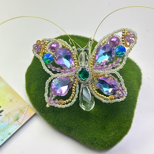 May include: A decorative butterfly brooch, featuring iridescent purple, blue, and green gemstones, and gold-coloured wire and beads. The butterfly is displayed on a green, textured surface. A card with the words "Thank You" is also visible.