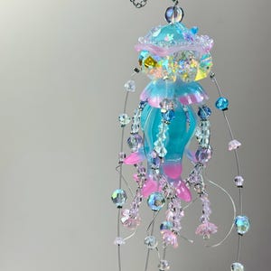 May include: A whimsical, handmade jellyfish mobile. It features a turquoise body with pink and blue accents, adorned with iridescent beads and delicate floral embellishments. The mobile is suspended by thin wires.