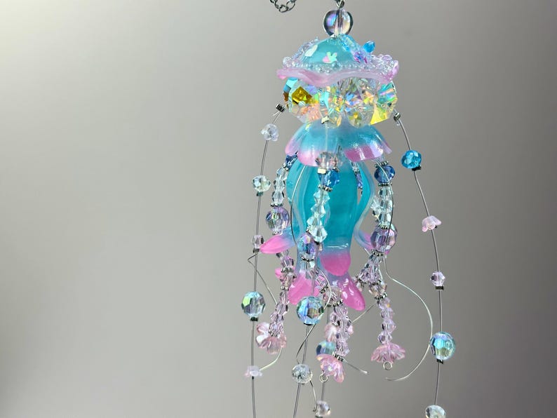 May include: A whimsical, handmade jellyfish mobile. It features a turquoise body with pink and blue accents, adorned with iridescent beads and delicate floral embellishments. The mobile is suspended by thin wires.