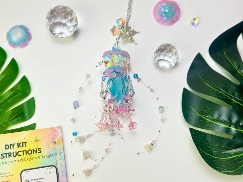 May include: A handmade jellyfish-shaped crystal suncatcher with a blue and pink color scheme. The suncatcher is adorned with beads and hanging strands. The image also includes decorative crystal balls, a monstera leaf, and a DIY kit instruction card.