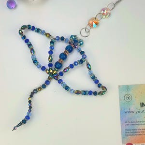 May include: A blue beaded suncatcher DIY kit with various shades of blue beads and iridescent accents. The kit includes instructions and a tutorial link. Additional iridescent crystals and a decorative chain are also visible.