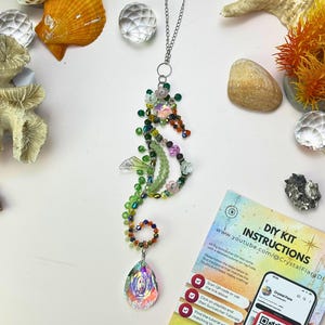 May include: A beaded seahorse pendant necklace with a silver chain. The seahorse is made of green, orange, and pink beads. A teardrop-shaped iridescent crystal hangs below. The image includes seashells, crystals, and a DIY kit instruction sheet.