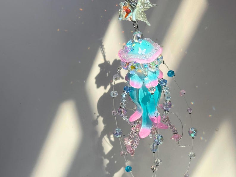 May include: A whimsical jellyfish-shaped suncatcher. The suncatcher features a translucent blue and pink jellyfish design with beaded accents. The jellyfish has a pink and blue top with a butterfly design. The suncatcher is suspended by a silver chain.