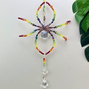 May include: A colorful beaded spider suncatcher with a large crystal ball body and teardrop pendant. The spider's legs and body are made of various colored beads, including red, orange, yellow, green, and purple. The suncatcher is designed to hang and catch the light.