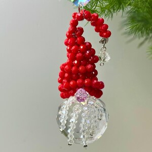 Könnte beinhalten: Festlicher Schmuck mit roter Perlenmütze und einer durchsichtigen Kristallkugel mit hängenden Kristallsträngen. Ein kleiner, hängender Kristallakzent unterstreicht das Design. Der Schmuck hängt vor einem sanften grünen Hintergrund.