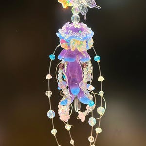 May include: A decorative hanging ornament featuring a jellyfish design. The jellyfish is purple with blue accents and adorned with iridescent flowers and beads. A clear butterfly and flowers are at the top, with beaded strands hanging below.
