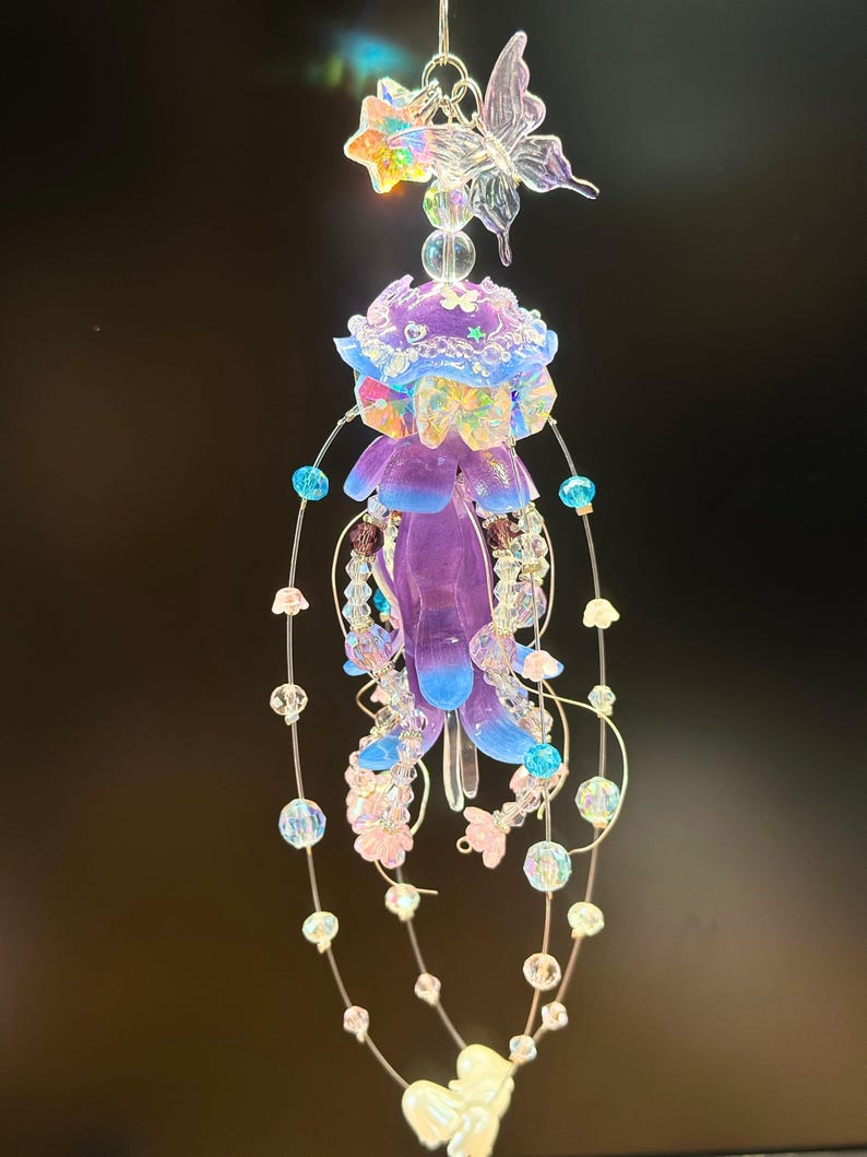 May include: A decorative hanging ornament featuring a jellyfish design. The jellyfish is purple with blue accents and adorned with iridescent flowers and beads. A clear butterfly and flowers are at the top, with beaded strands hanging below.