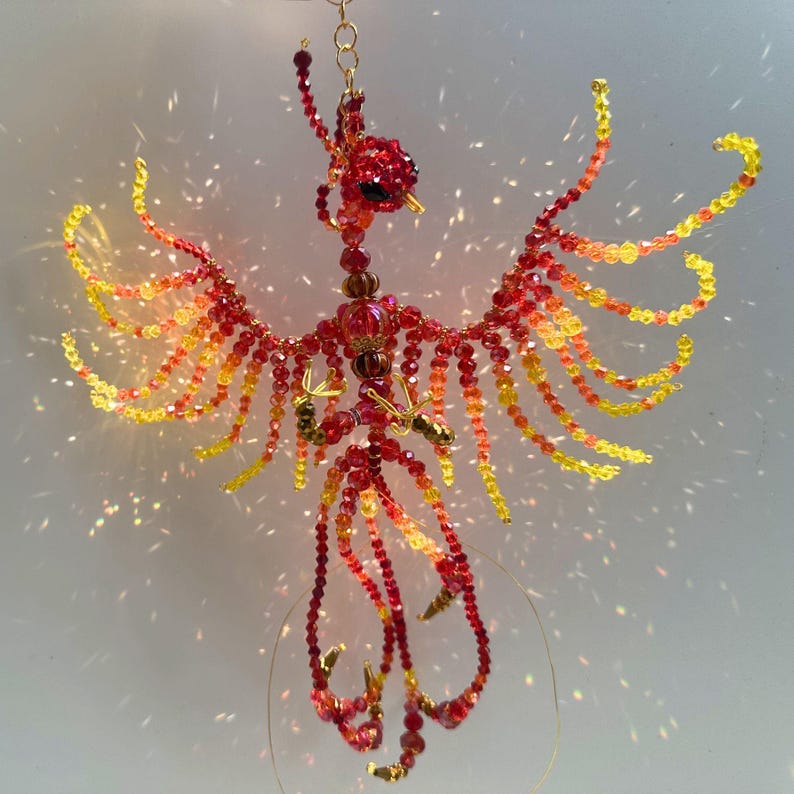May include: A handcrafted beaded phoenix ornament. The bird features a red, orange, and yellow color scheme, with a gold-colored wire frame. The wings are spread, and the bird is suspended from a gold chain. The ornament is designed to catch the light.