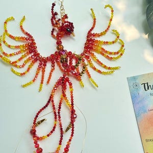 May include: A handcrafted phoenix sculpture made of red, orange, and yellow beads. The beaded bird has outstretched wings and a long tail, with a small head and beak. The sculpture is on a white surface. A card with the text "Thank you" is in the background.