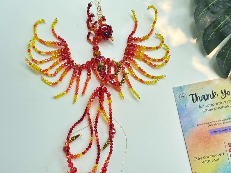 May include: A handcrafted phoenix sculpture made of red, orange, and yellow beads. The beaded bird has outstretched wings and a long tail, with a small head and beak. The sculpture is on a white surface. A card with the text "Thank you" is in the background.