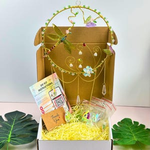 Könnte beinhalten: Ein DIY-Kit für einen dekorativen Traumfänger. Der goldfarbene Metallrahmen ist mit einer Libelle, Schmetterlingen und hängenden Kristallen verziert. Das Set enthält Anweisungen und eine Dankeskarte, alles in einem Karton mit gelber Füllung.
