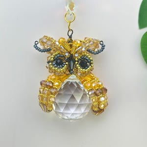 May include: A handcrafted owl ornament made of gold and amber-coloured beads, with dark blue accents. The owl features a clear crystal ball as its body and is suspended by a gold chain. The owl's eyes are black. The ornament is designed for decoration.