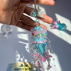 May include: A handmade jellyfish pendant with a chain. The jellyfish features a blue and pink body with dangling beaded strands. A small, iridescent butterfly and crystal accents are attached to the chain. The DIY kit instructions are visible in the background.