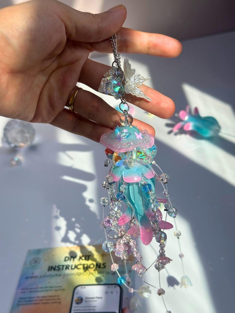 May include: A handmade jellyfish pendant with a chain. The jellyfish features a blue and pink body with dangling beaded strands. A small, iridescent butterfly and crystal accents are attached to the chain. The DIY kit instructions are visible in the background.