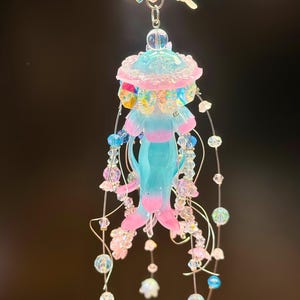 May include: A whimsical, handmade jellyfish ornament. The ornament features a light blue body with pink and blue accents, adorned with iridescent beads and dangling floral charms. A fairy-shaped charm and star-shaped charm are at the top.